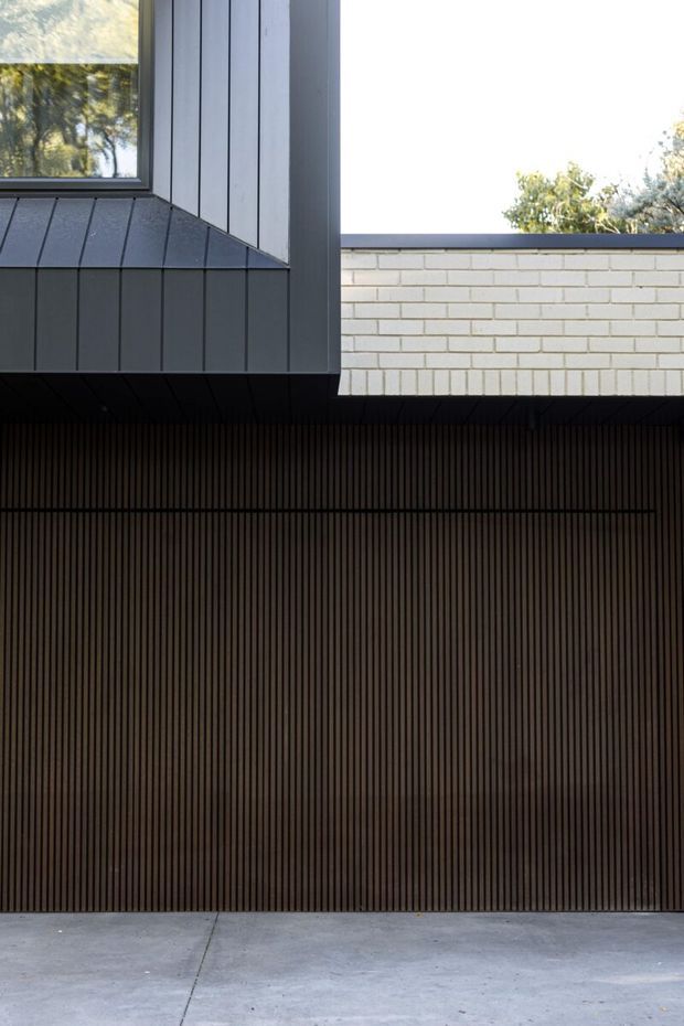 A close-up exterior architectural view showing a dark, vertically ribbed garage door or screened facade beneath a projecting upper storey. The upper level combines charcoal vertical metal cladding, a dark-framed window, and a pale rendered or painted brick wall with a dark parapet edge. A concrete driveway occupies the foreground, with trees visible beyond the roofline. The composition is minimalist and strongly geometric.