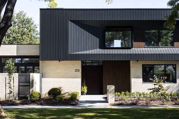Front exterior view of a contemporary two-storey residence with pale brick walls, dark vertical metal cladding, a projecting upper level, black-framed windows, and a recessed timber-toned entry. Low landscaped planting and a lawn sit in front of the house.