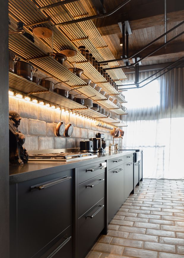 A narrow, richly detailed industrial-style kitchen with a long run of dark matte cabinetry and drawers, dark benchtops, integrated cooking appliances, and open shelving suspended above the work zone. The shelves display cookware and crockery, while pots hang from the backsplash. A large bright curtained window illuminates the space. The ceiling combines exposed dark timber and structural framing with slatted timber shelving/ceiling elements, and the floor is finished in rectangular rustic brick-toned tiles.
