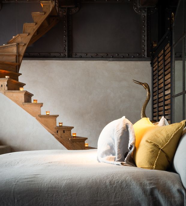 A moody, industrial-style living room dominated by a large upholstered sofa in pale grey or off-white, layered with white and mustard cushions. A timber staircase rises along the left wall, with small candles placed on the steps. The room combines textured plaster walls, dark riveted metal wall panelling, black-framed glazing or joinery, and a sculptural decorative object, creating a warm and dramatic atmosphere.