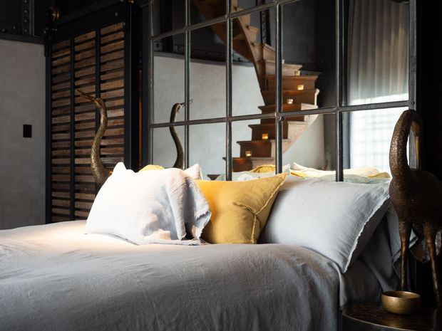 A moody, refined bedroom with a neatly made bed dressed in white and pale blue linens, accented by a mustard-yellow cushion. Behind the bed is a large black metal-framed mirrored or glazed partition reflecting a timber staircase, alongside dark timber slatted joinery and sculptural dark-toned decor. The room combines industrial metal detailing with warm timber elements and soft textiles.