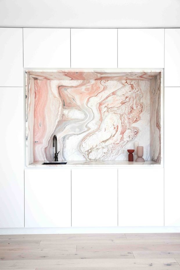 A minimalist white kitchen wall with a recessed open niche framed in pale pink-and-white veined natural stone. The niche contains a black gooseneck tap and basin at the lower left, with small decorative vessels at the right. Pale timber flooring runs across the foreground.