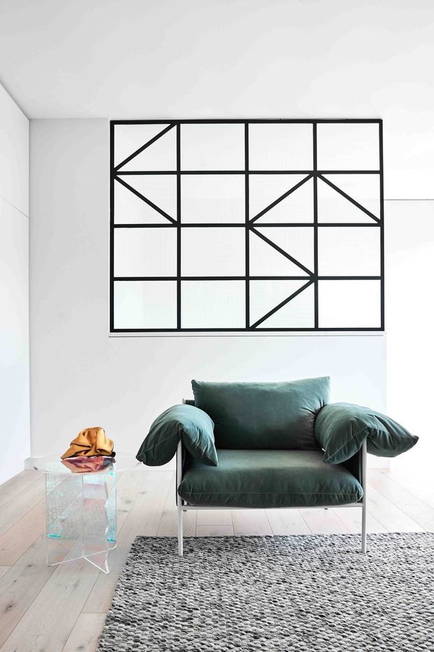 Bright minimalist living room featuring a deep green upholstered lounge chair with slim metal legs, a transparent glass side table, pale timber flooring, and a large grey textured rug. A bold black geometric grid artwork dominates the white wall behind the chair.