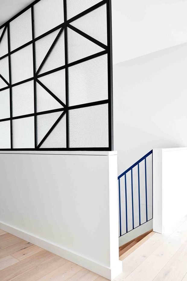 Bright minimalist interior featuring a stairwell and landing with pale timber flooring, white walls, and distinctive geometric metal balustrade screens. The upper balustrade uses black framing with translucent ribbed infill and triangular bracing, while the stair opening below has a deep blue metal railing.