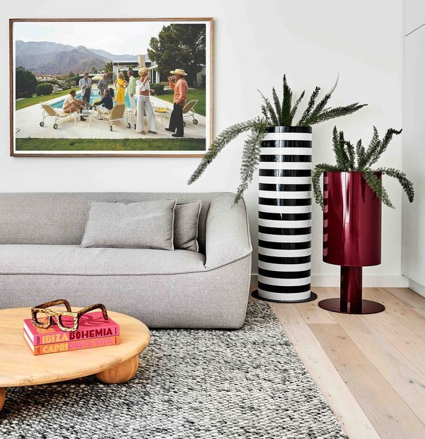 A bright contemporary living room with a light grey upholstered sofa, pale timber flooring, a textured grey rug, and a rounded light-wood coffee table. A striking black-and-white striped cylindrical planter and deep red sculptural floor lamp add bold accents, while framed artwork hangs above the sofa.