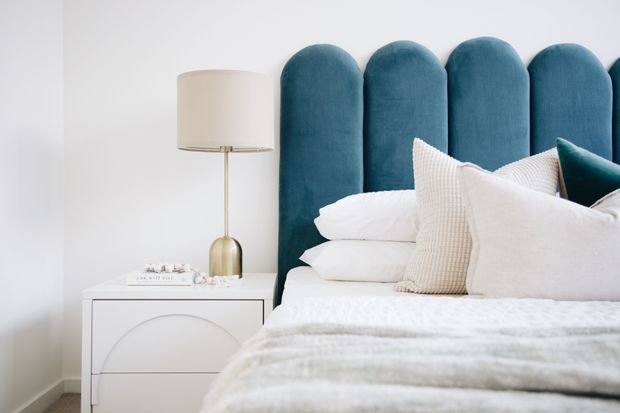 Bright, refined bedroom interior featuring a tall scalloped deep teal upholstered headboard, layered white bedding, textured cream and white pillows, and a white curved-front bedside table with a brass-toned lamp. The palette is soft and airy with a sophisticated contemporary feel.