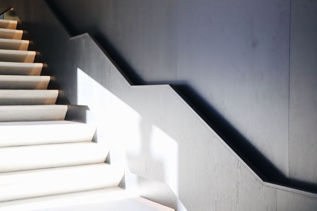 Close-up architectural view of a modern stairway with pale timber or concrete-toned treads, dark charcoal walls, and a sharply angled balustrade or stair-side wall. Strong sunlight creates geometric shadows across the steps and wall, emphasizing the minimalist composition.
