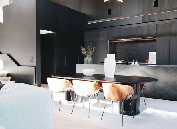 A sophisticated open-plan kitchen and dining area with dramatic dark cabinetry and wall panelling, a large stone island with integrated sink and black tapware, and a round black dining table with upholstered chairs. Bright natural light contrasts with the charcoal finishes, while a partial living area and staircase are visible at left.