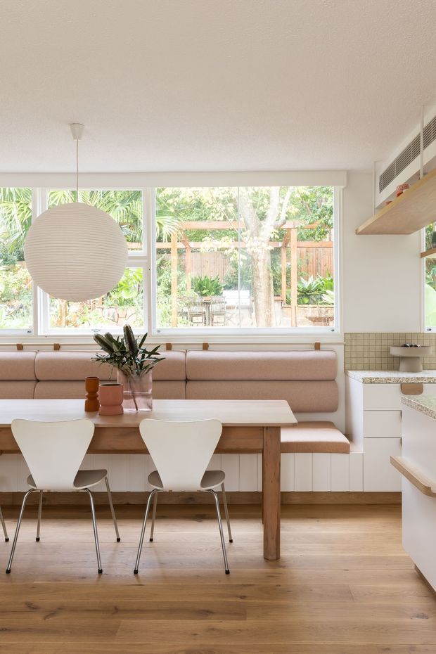 Bright, warmly styled open-plan kitchen and dining nook. A built-in blush upholstered banquette sits beneath large windows, paired with a light timber dining table and sculptural white chairs. The kitchen has white cabinetry, pale stone benchtops, a small square-tile splashback, open timber shelving, and integrated upper ventilation. A large spherical pendant light and indoor plant add soft decorative accents, while the timber flooring creates a cohesive natural base.