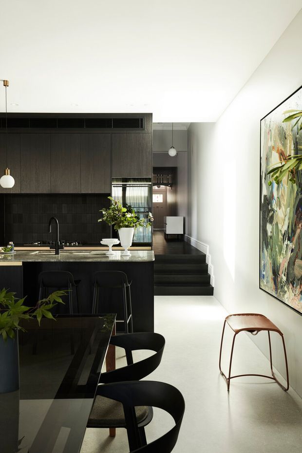 A refined open-plan kitchen and dining area with dark timber cabinetry, a black tiled splashback, stone benchtop and a black island with integrated sink. The space opens toward a short set of dark stairs and a hallway, with a large abstract artwork on the white wall. Black dining chairs, a dark glossy dining table, pendant lights and restrained greenery create a sophisticated, high-contrast interior.