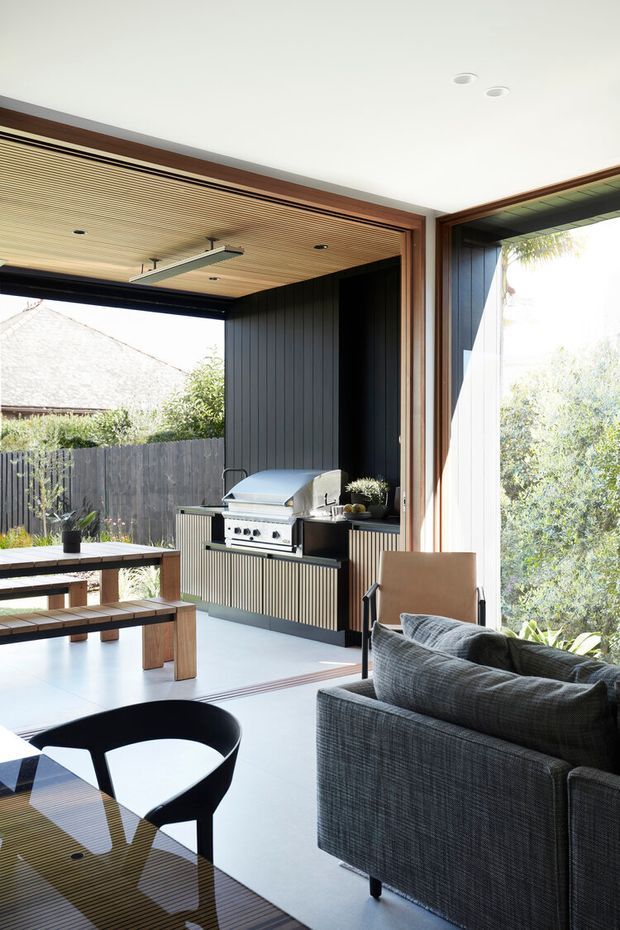 A bright, contemporary indoor-outdoor entertaining space with a covered barbecue kitchen opening onto a garden. The room features a timber-slatted ceiling, dark vertical wall cladding, large glazed openings, a built-in stainless-steel grill, timber picnic-style dining table, and dark upholstered lounge seating.