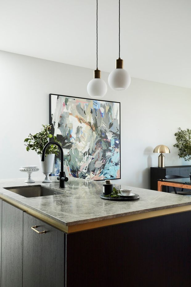A refined contemporary kitchen focused on a large island with a grey veined stone benchtop, integrated sink, and matte black gooseneck tap. The island has dark vertical-grain cabinetry with a contrasting metallic trim. Two globe pendant lights hang above, while an abstract artwork, sculptural table lamp, plants, and serving pieces add decorative character.