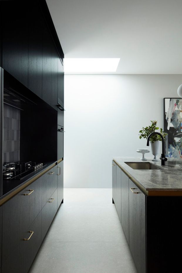 A narrow, contemporary galley kitchen with tall dark timber cabinetry along the left wall and a central island on the right. The island has a grey stone benchtop, integrated sink, black curved tapware, and dark cabinetry with brass handles. A gas cooktop and dark splashback are visible on the left, while a skylight brings bright natural light into the otherwise restrained space. Artwork and a potted plant add visual interest.