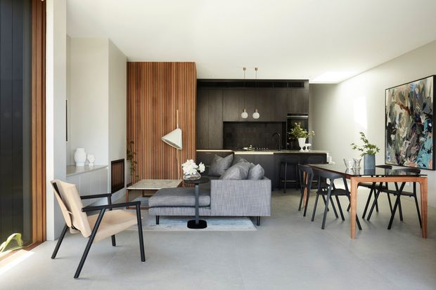 Open-plan contemporary living and dining area with a compact black kitchen at the rear. The room features a grey sectional sofa, timber and black dining furniture, a vertical timber slat screen, pale concrete-look flooring, light walls, a large abstract artwork, and abundant natural light.