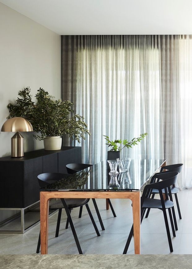 A refined dining area with a black glass-topped dining table and sculptural black chairs, positioned in front of full-height sheer curtains. A dark built-in sideboard with indoor plants and a brass-toned dome lamp lines the wall. The room has pale walls, a polished light grey floor, and a restrained, contemporary palette with strong natural light.