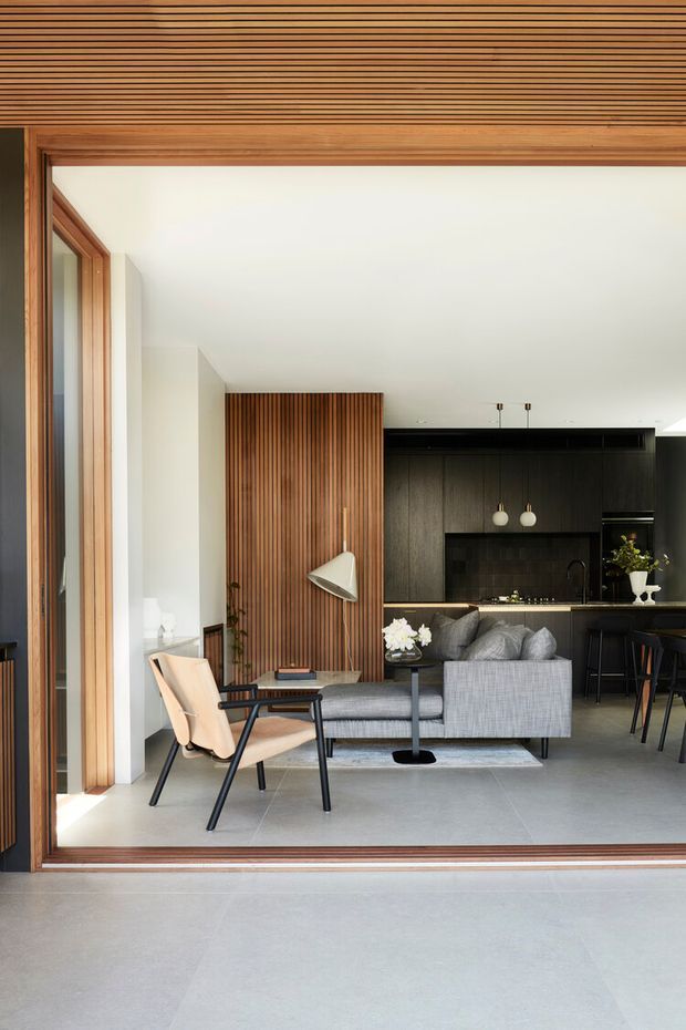 A refined open-plan living space viewed through a large timber-framed opening. The room combines a grey sofa, lounge chair, small side tables and a floor lamp with a dark contemporary kitchen at the rear. Vertical timber slat panelling forms a feature wall, while the ceiling has fine linear timber battens and the floor is a large-format light grey finish. The space is bright, calm and connected to an exterior threshold.