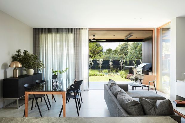 Bright contemporary open-plan living and dining area opening onto a landscaped outdoor space. The room features a grey sectional sofa, timber dining table with dark chairs, sheer full-height curtains, black built-in cabinetry, a ceiling fan, and large glazed openings. A covered outdoor entertaining area with barbecue is visible beyond.