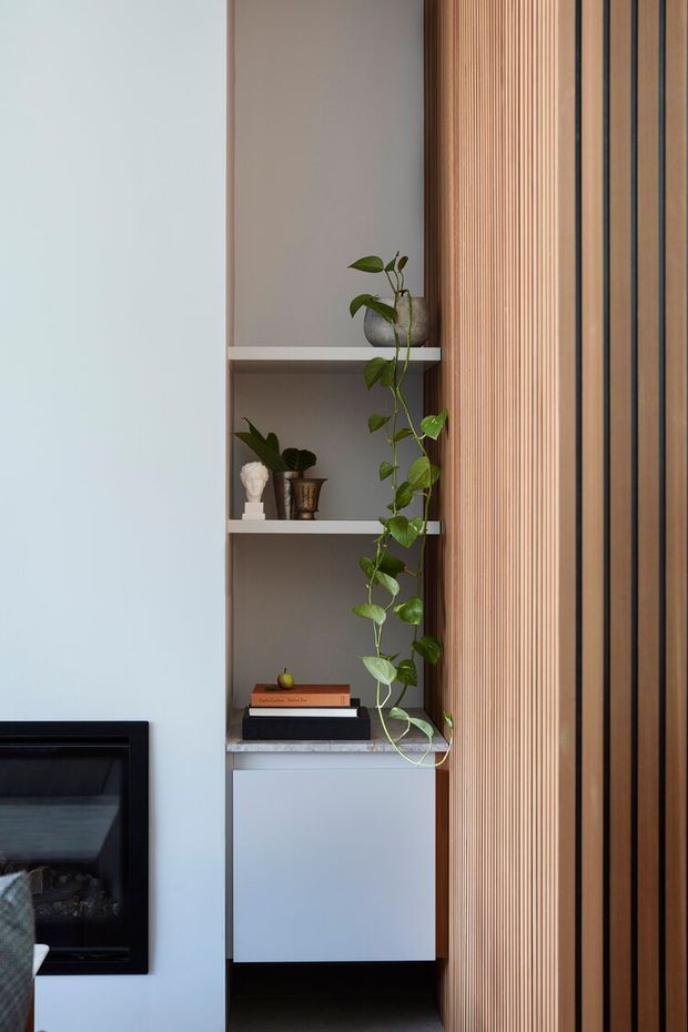 A close-up interior detail showing a built-in shelving niche with grey shelving, books, small decorative objects, and a trailing indoor plant. A black fireplace is partially visible at left, while a prominent vertical timber slatted wall or screen occupies the right side. The palette is restrained and contemporary, with pale walls, light timber, and dark accents.
