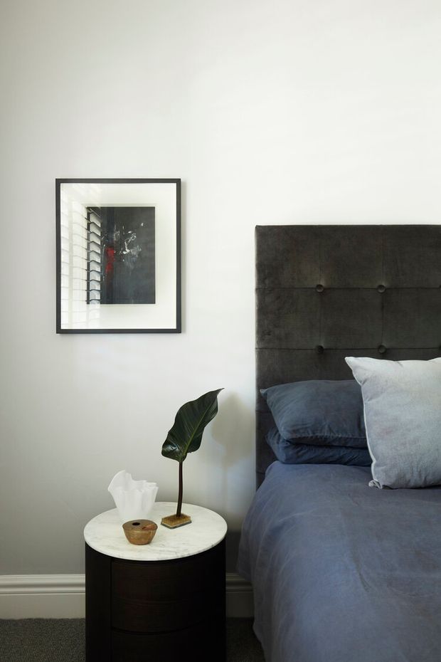 A close-up view of a refined bedroom corner featuring a dark upholstered, button-tufted headboard, blue-grey bedding, and a round dark timber bedside table topped with a white marble surface. A simple framed artwork hangs on the pale wall, with a small potted tropical leaf and tissue holder adding understated detail. The room has a calm, moody and contemporary feel.