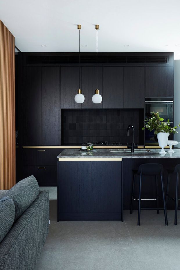 A refined open-plan kitchen viewed from the adjacent living area, featuring dark timber or black cabinetry, a black tiled splashback, a grey stone island benchtop, integrated appliances, brass pendant details, and a black kitchen faucet. A grey sofa is partially visible in the foreground, with bar seating at the island. Vertical timber battens add warmth along the left wall.