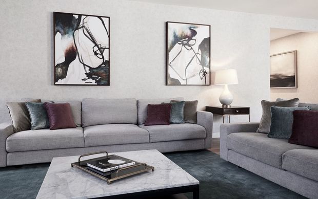 A refined contemporary living room with a large grey sectional sofa and matching armchair, arranged around a rectangular marble coffee table. The room features soft neutral walls, dark blue-grey carpeting, abstract monochromatic artwork, and a small dark timber side table with a ceramic lamp. Jewel-toned cushions add subtle burgundy and blue accents.