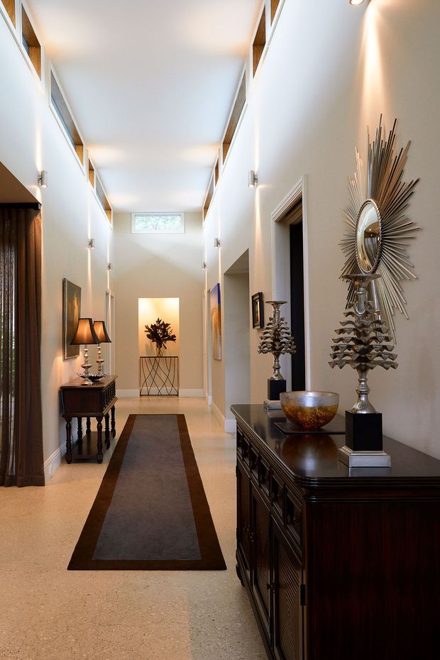A tall, refined residential hallway with a long central runner, pale walls and floor, high ceiling, clerestory-style glazing, dark timber console furniture, lamps, decorative candlesticks, and a prominent sunburst mirror. Multiple doorways and openings line the corridor, creating a formal gallery-like entry passage.