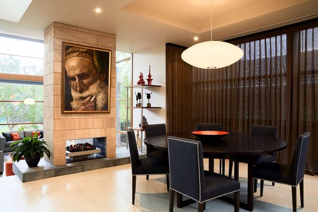 A refined open-plan dining and living area with a dark round dining table and upholstered dining chairs positioned beside a timber-clad fireplace feature wall. The fireplace surround includes a framed artwork above a linear firebox, while floor-to-ceiling glazing with dark sheer curtains spans the rear wall. Warm recessed and pendant lighting, polished light flooring, shelving, and indoor greenery create an elegant, layered interior.