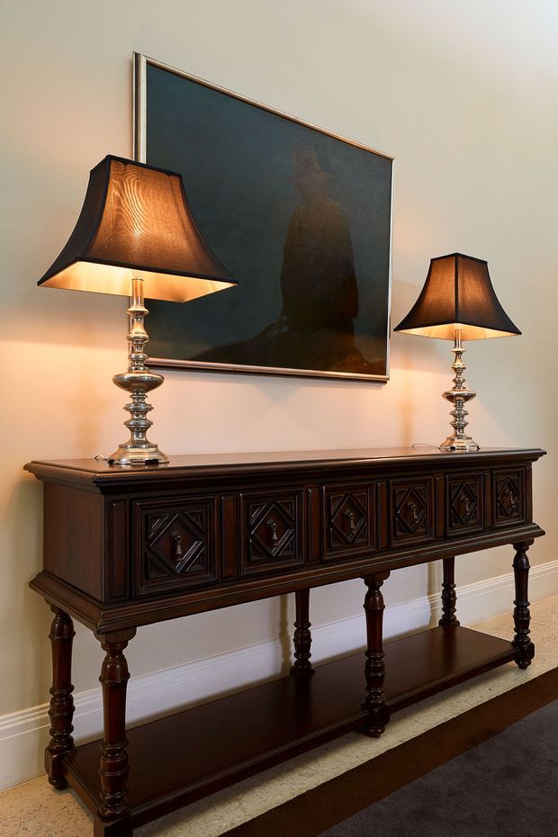 A refined interior vignette featuring a dark, ornately carved wooden console table with turned legs and paneled drawer fronts. Two matching traditional table lamps with dark fabric shades sit on the console, beneath a large dark-toned framed artwork or mirror. The setting has warm neutral walls, white skirting, and a layered neutral rug, creating a formal, classic atmosphere.