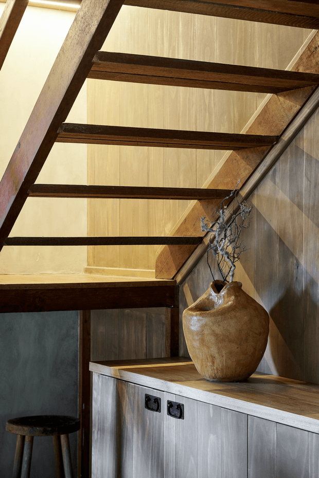 Interior architectural detail showing a timber stair or loft structure with exposed dark wood beams and warm translucent vertical-panel walls. A built-in grey timber cabinet with a wood benchtop sits beneath the stairs, styled with a large rustic ceramic vessel and dried branches. The composition has a warm, tactile, rustic-industrial character.