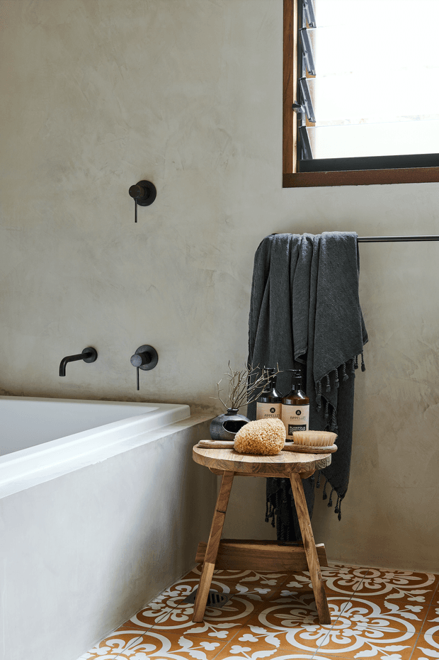 A warmly styled bathroom featuring a white built-in bathtub with dark wall-mounted fixtures, a textured warm beige plaster wall, timber-framed louvred window, and dark grey towels on a black rail. A small rustic timber stool holds natural bath accessories and amber bottles. Decorative brown, cream, and terracotta patterned floor tiles add a strong feature element.