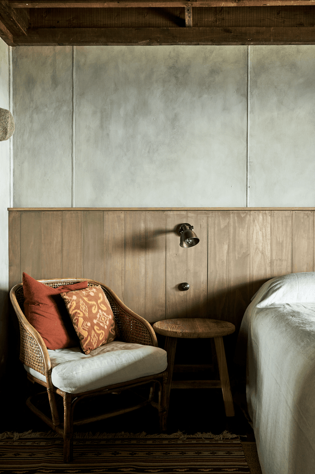 A warmly lit rustic bedroom corner featuring a cane armchair with patterned terracotta cushions, a small round wooden stool, and the edge of a bed dressed in white linen. Vertical timber wall panelling, a pale textured plaster upper wall, exposed dark timber ceiling beams, a wall-mounted reading light, and a patterned rug create an earthy, tactile interior.