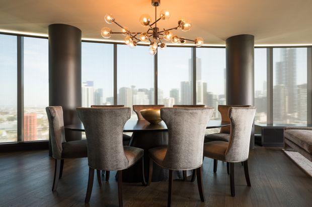 A sophisticated high-rise dining area with a large dark wood dining table surrounded by upholstered dining chairs. Floor-to-ceiling curved glazing provides an expansive urban skyline view, while cylindrical structural columns, dark timber flooring, and a sculptural branching pendant light create a refined contemporary interior.