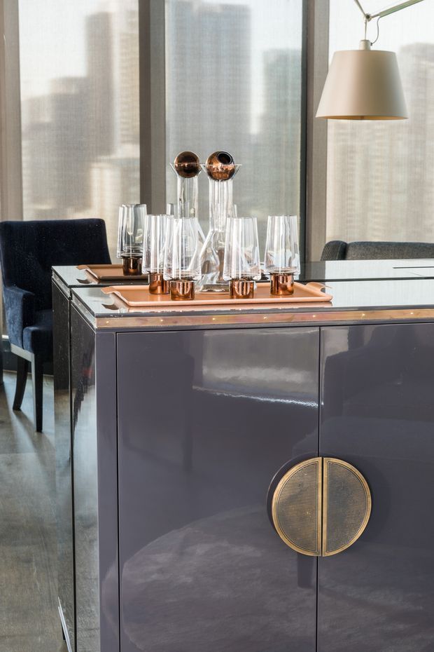 A refined dining area centered on a glossy dark charcoal sideboard or buffet with large circular brass-gold handles. The sideboard is styled with copper-toned trays, glassware, decanters, and sculptural vessels. Dark upholstered dining chairs, sheer floor-to-ceiling window coverings, and a modern brass-and-cream pendant light complete the sophisticated interior.