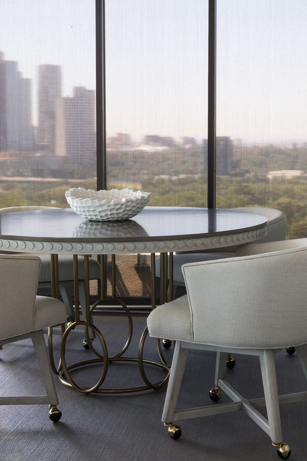 Elegant dining area with a round reflective dining table featuring an ornate bronze-toned circular base, upholstered light-grey dining chairs, and a sculptural white bowl centerpiece. Floor-to-ceiling windows with sheer blinds overlook a distant urban skyline and greenery.