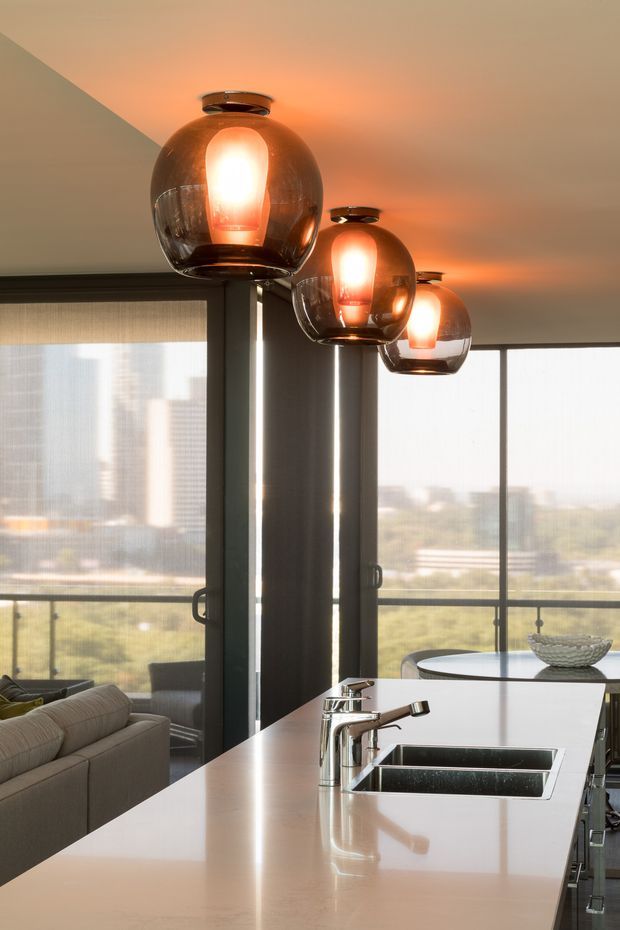 A contemporary open-plan kitchen with a large pale stone island, integrated double stainless-steel sink and chrome tapware. Three smoked-glass pendant lights with glowing warm bulbs hang above the island. Floor-to-ceiling glazing and sliding doors open toward a balcony or exterior view, with a sofa and dining area visible in the background.