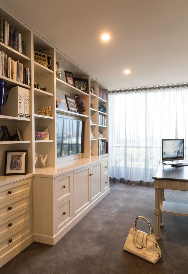 A bright, elegant home office with extensive custom built-in bookcases and lower cabinetry along one wall. The shelving displays books, framed photographs, ornaments and a television set into the joinery. A traditional-style desk with a computer sits beside a large window dressed with sheer curtains. The room has recessed ceiling lights, warm cream cabinetry and grey carpet, creating a refined, comfortable workspace.