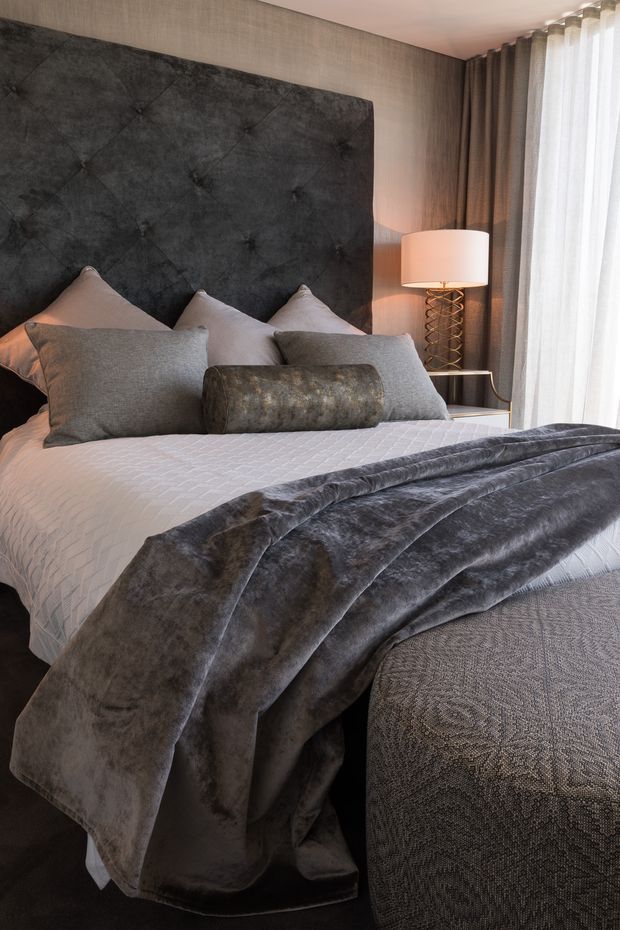 A luxurious bedroom interior centred on a neatly styled bed with a tall charcoal-grey tufted upholstered headboard, layered grey and taupe pillows, a white quilted coverlet, and a plush dark grey throw. Floor-to-ceiling sheer and heavy curtains frame the window, while a sculptural bedside lamp with a metallic spiral base adds a refined accent. The palette is soft, muted, and monochromatic with tactile fabrics throughout.