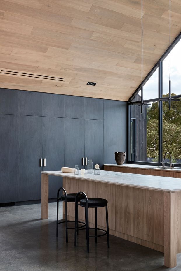 A contemporary kitchen with a large timber island, pale stone benchtop, dark full-height cabinetry, polished concrete flooring, and a dramatic sloped timber-lined ceiling. Black-framed floor-to-ceiling glazing brings in views of trees, while black bar stools and minimal pendant lighting add contrast.