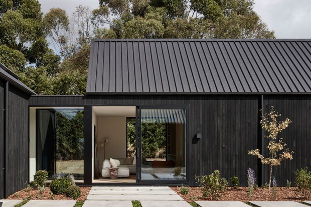 Exterior view of a compact, contemporary single-storey dwelling clad in dark vertical timber boards. A steep simple gable roof with dark standing-seam metal roofing dominates the composition, with full-height glazed sliding doors opening to a light interior. A restrained landscaped entry path and native vegetation surround the building.