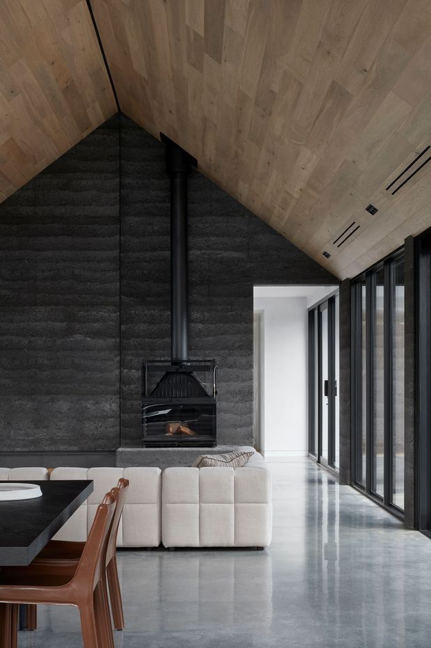 A dramatic contemporary living room with a steeply pitched timber-lined ceiling, dark textured feature wall and black freestanding wood-burning fireplace. A pale modular sofa faces the fireplace, while full-height black-framed glazing runs along the right side, creating a bright, elongated space. A dark dining table and brown leather dining chairs are partially visible in the foreground. The polished grey concrete floor reflects the glazing and furniture.
