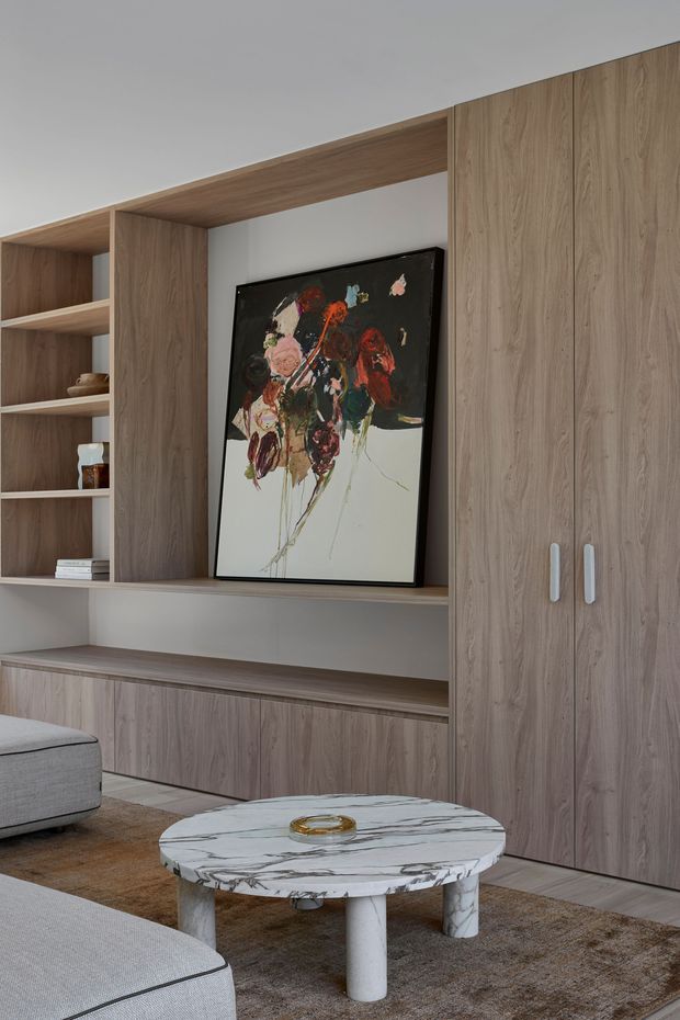 A refined living room featuring a full-height timber-look built-in entertainment and storage wall with open shelving, lower cabinetry, and tall cupboard doors. A large abstract artwork is displayed in the central niche. Partial views of a light grey sectional sofa, round marble coffee table, and neutral textured rug create a calm, contemporary interior.