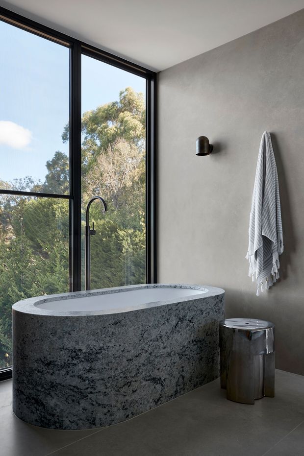 A serene contemporary bathroom featuring a freestanding oval bathtub with a dark, heavily textured stone exterior and pale stone-like interior. Full-height black-framed glazing overlooks trees, while a grey plastered wall has a minimalist black wall light and a hanging textured towel. A small metallic stool sits beside the bath, and large dark grey floor tiles complete the restrained, nature-connected scheme.