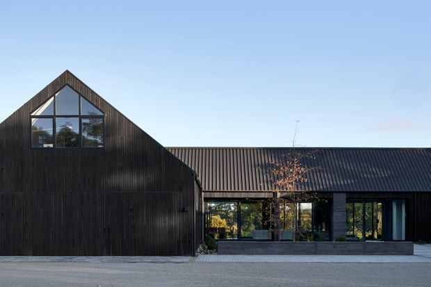 Exterior view of a contemporary single-storey building composed of a steep gabled wing and a long lower pitched-roof wing. The facade is clad in dark vertical timber boards, with large black-framed glazed openings and a recessed central entry. A paved forecourt sits in front of the building, with minimal landscaping and clear blue sky.