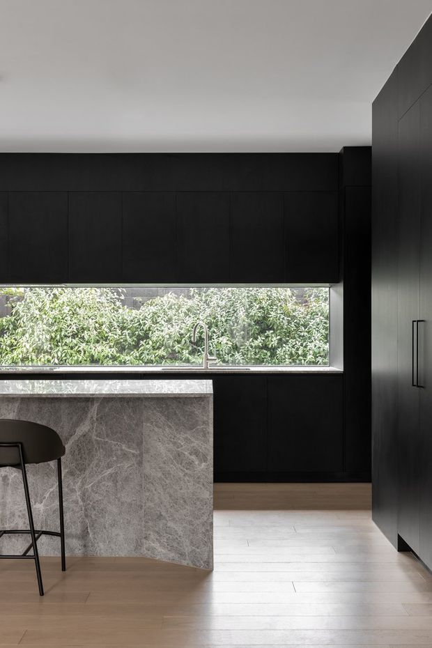 A refined contemporary kitchen with extensive matte black cabinetry, a large grey-veined stone island and benchtop, integrated sink and faucet, and a long horizontal window overlooking dense greenery. Light timber flooring contrasts with the dark joinery, while a partial bar stool is visible at the island.