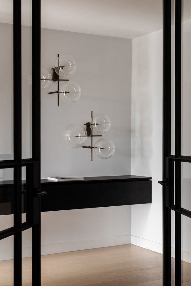 A minimalist interior vignette viewed through black-framed glazed doors, featuring a floating black console with a dark stone-like top, sculptural globe pendant lights, pale timber flooring, and white walls. The space is clean, refined, and softly lit.