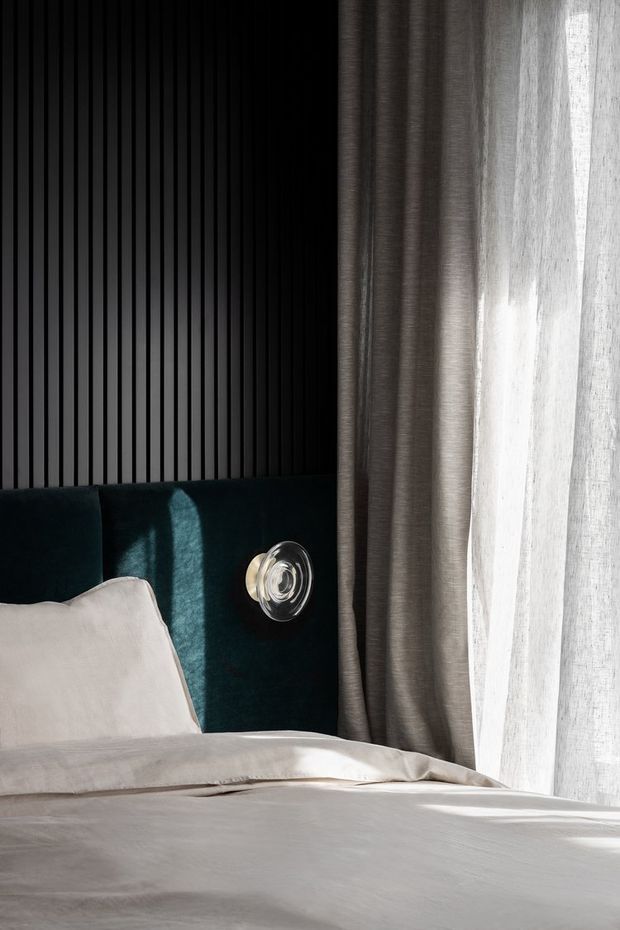 Close-up of a refined bedroom corner featuring a bed with white bedding, a deep teal upholstered headboard, a black vertical slatted feature wall, a wall-mounted round reading light, and layered grey and sheer curtains beside a bright window. The image has strong natural light and a high-contrast, contemporary feel.