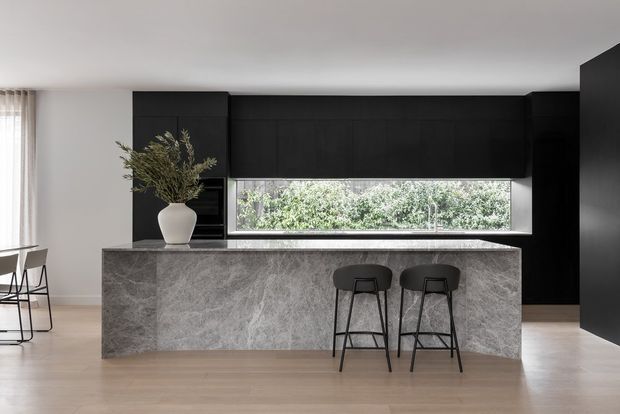Contemporary kitchen featuring a large grey stone island with waterfall-style sides, two black bar stools, extensive matte-black cabinetry, a long horizontal window, pale timber flooring, and restrained neutral styling. A small dining area is partially visible at the left.