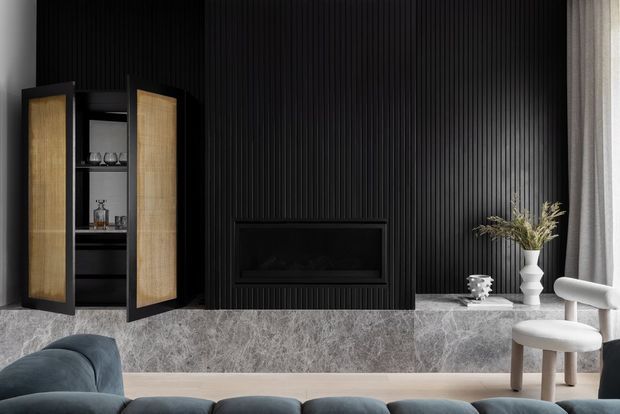 A sophisticated contemporary living room featuring a dramatic black vertical-ribbed feature wall with a built-in fireplace, flanked by a tall timber-fronted bar or display cabinet. A grey marble or stone hearth extends across the wall, styled with a vase and decor. Soft grey seating and a sculptural white lounge chair sit in front of the fireplace, with sheer curtains bringing in natural light.