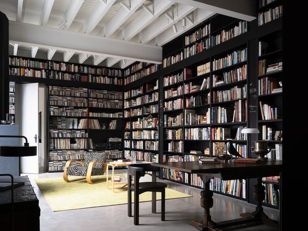 A sophisticated library and study with floor-to-ceiling black built-in bookshelves filled with books, a large dark wood writing table, chairs, and a small reading area. White exposed roof trusses and ceiling beams contrast with the dark joinery, while polished flooring and abundant daylight create a refined, dramatic atmosphere.