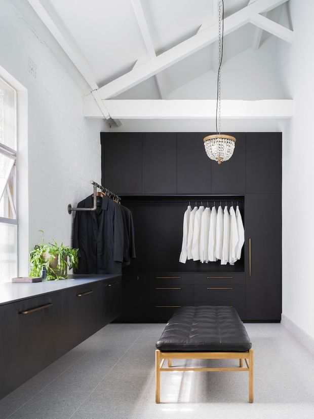 Bright, refined walk-in wardrobe with extensive matte black built-in cabinetry, open hanging rails, drawers, and a central black upholstered bench with a timber-toned frame. White walls and exposed painted roof trusses create a high, airy ceiling, while a large window provides natural light. A decorative pendant and potted plants add contrast.